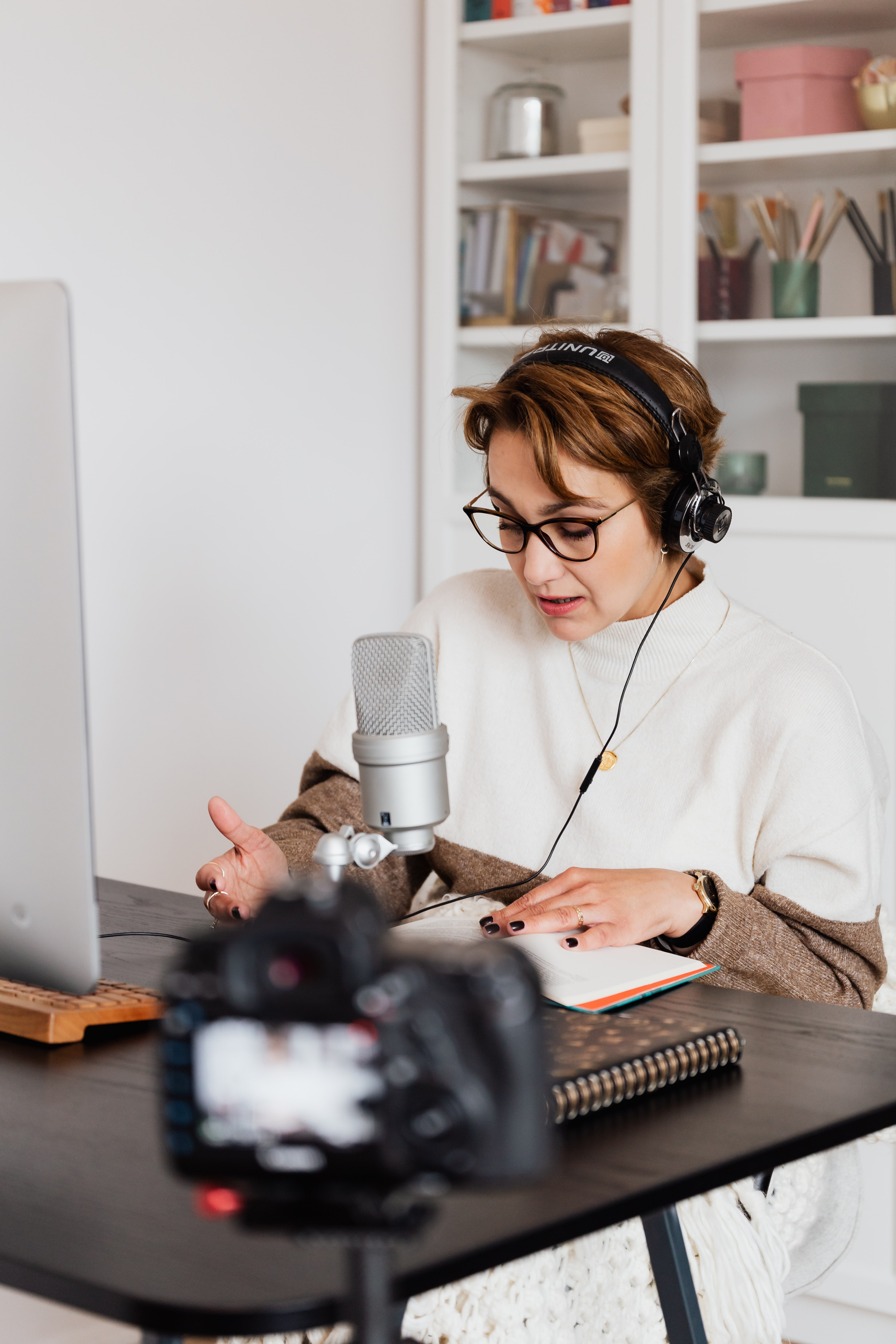 A woman sits at a desk and speaks into a microphone, being recorded by a camera.