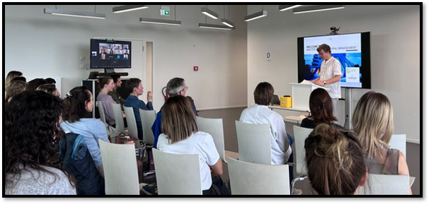 Die Abbildung zeigt das Setting aus der Sicht einer hinteren Tischreihe von rechts. Wir sehen vorne einen Dozenten mit der gelben Catchbox sowie links die Online-Teilnehmenden auf einem Monitor. Im Hintergrund ist eine Präsentation auf einem Display geteilt.