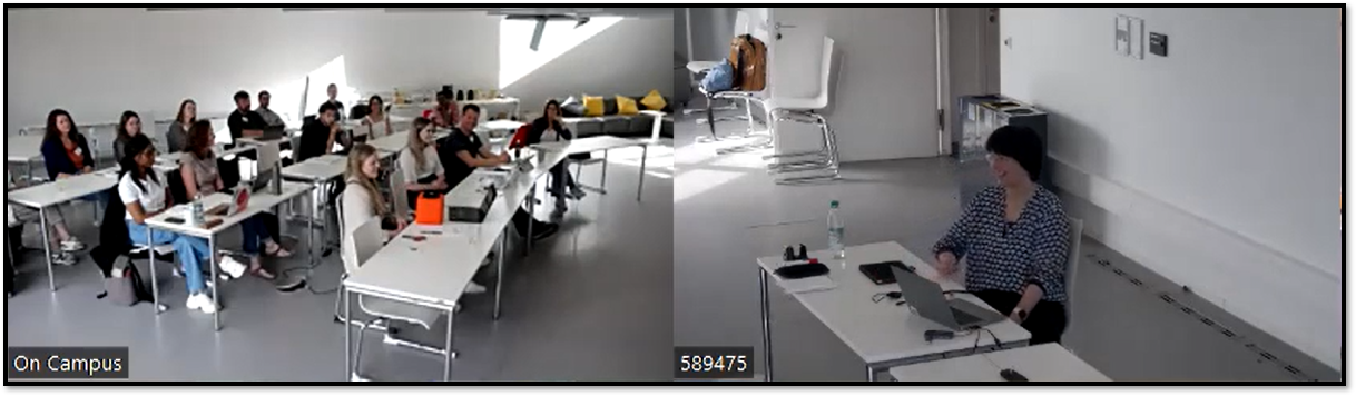 Die Abbildung zeigt den View der Online-Teilnehmenden über die beiden Kameras in den Präsenzraum mithilfe zweier Zoom-Kacheln. Rechts ist die Dozentin auf einem Stuhl sitzend groß abgebildet, links sind die Tischreihen mitsamt der Studierenden vor Ort zu sehen.