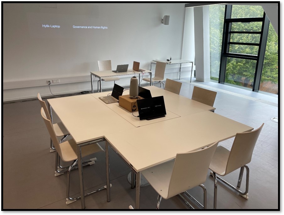 Die Abbildung zeigt die reale Umsetzung der Grafik: Eine Tischinsel mit Meeting Owl und Zusatzbildschirmen in der Mitte. Sechs Stühle stehen unbesetzt um die Insel herum. Im Hintergrund ist eine Videokonferenz auf einem Beamer zu sehen.