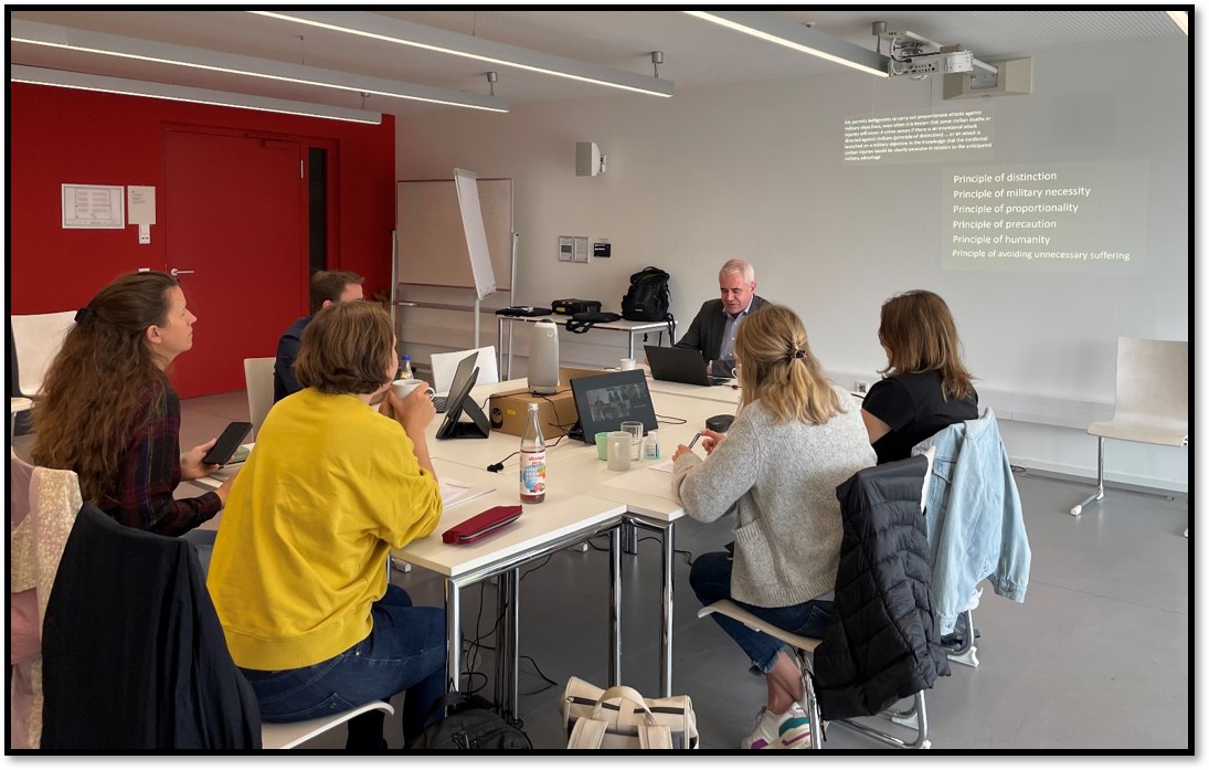 Die Abbildung zeigt eine Tischinsel mit Meeting Owl und Zusatzbildschirmen in der Mitte. Auf den Zusatzbildschirmen sind online Zugeschaltete zu sehen. Fünf Studierende sitzen um die Insel herum. Am vorderen Ende der Insel sitzt ein Dozent. Im Hintergrund ist eine in Zoom geteilte Präsentation zu sehen.