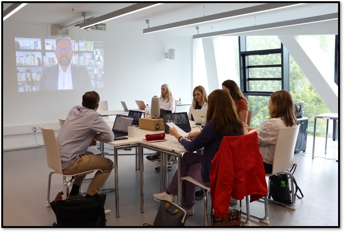 Die Abbildung zeigt eine Tischinsel mit Meeting Owl und Zusatzbildschirmen in der Mitte. Auf den Zusatzbildschirmen sind online Zugeschaltete zu sehen. Fünf Studierende sitzen um die Insel herum. Im Hintergrund ist der Dozierende online zugeschaltet auf dem Beamerbild zu sehen.